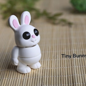 May include: A white, segmented, articulated toy bunny with black eyes and pink ears. The bunny is standing on a woven bamboo mat. The text "Tiny Bunny" is visible in the image.