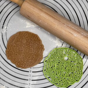 May include: A green plastic cookie cutter with the Hebrew words "Shavua Tov" (Happy Week) and a floral design. A brown cookie with the same design is shown next to it on a white and grey patterned surface.