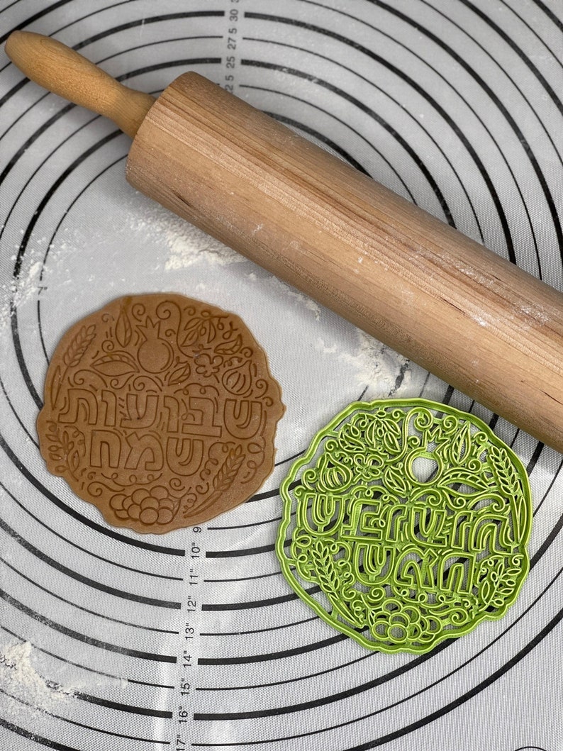 May include: A green plastic cookie cutter with the Hebrew words "Shavua Tov" (Happy Week) and a floral design. A brown cookie with the same design is shown next to it on a white and grey patterned surface.