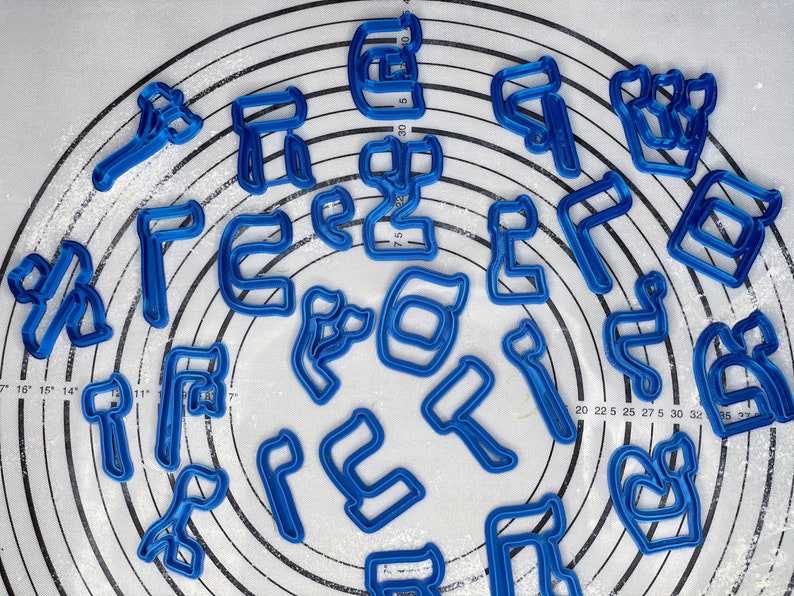 May include: A set of 20 blue plastic cookie cutters shaped like Hebrew letters. The cutters are arranged in a circle on a white surface with a ruler in the background.