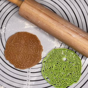 May include: Two cookie cutters on a white silicone baking mat with a rolling pin. The cookie cutters are round and feature a floral design with the Hebrew words "Shabbat Shalom" and "Happy Shabbat" in the center.