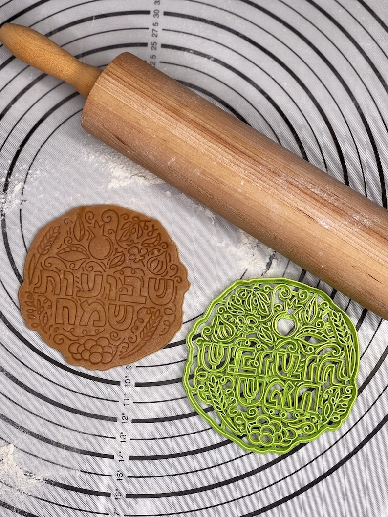 May include: Two cookie cutters on a white silicone baking mat with a rolling pin. The cookie cutters are round and feature a floral design with the Hebrew words "Shabbat Shalom" and "Happy Shabbat" in the center.