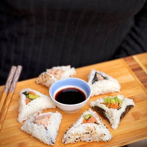 May include: A wooden tray with six triangular pieces of sushi, each with different fillings. A small white bowl with soy sauce is in the center of the tray.