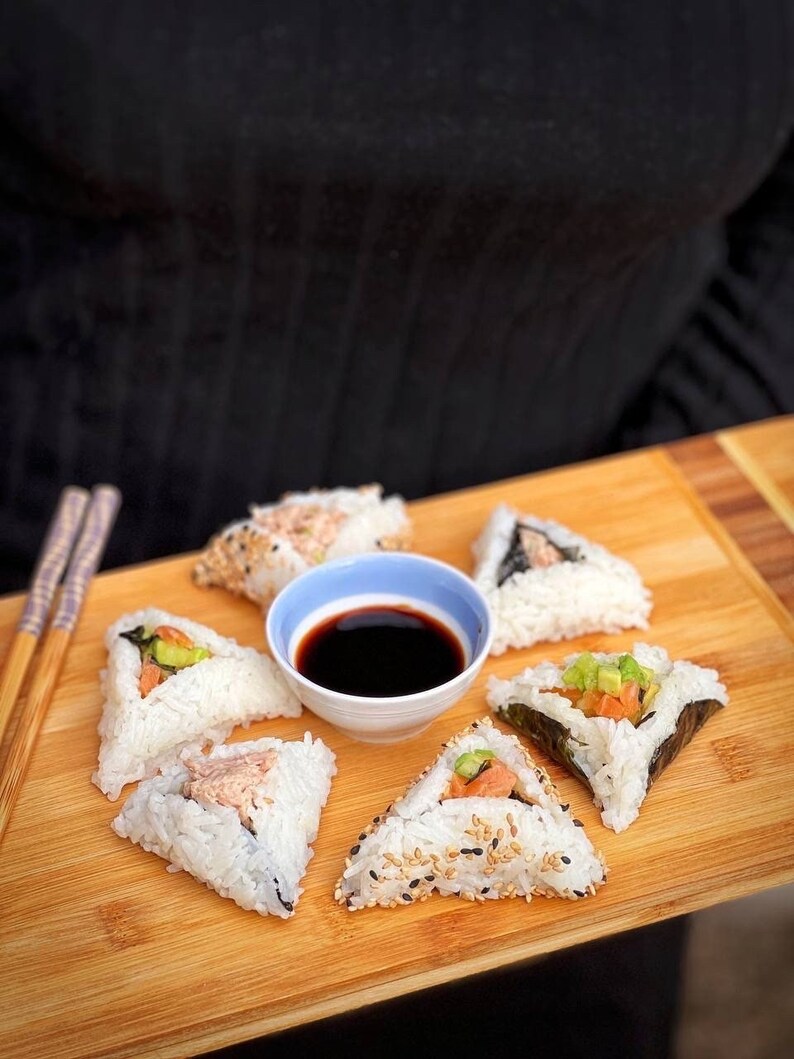 May include: A wooden tray with six triangular pieces of sushi, each with different fillings. A small white bowl with soy sauce is in the center of the tray.