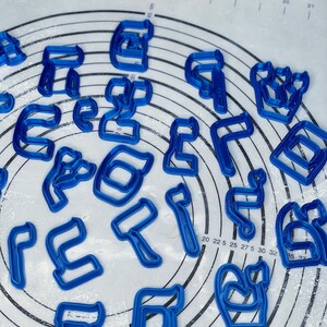 May include: A set of blue plastic cookie cutters shaped like Hebrew letters. The cutters are arranged in a circle on a white surface with a grid pattern.