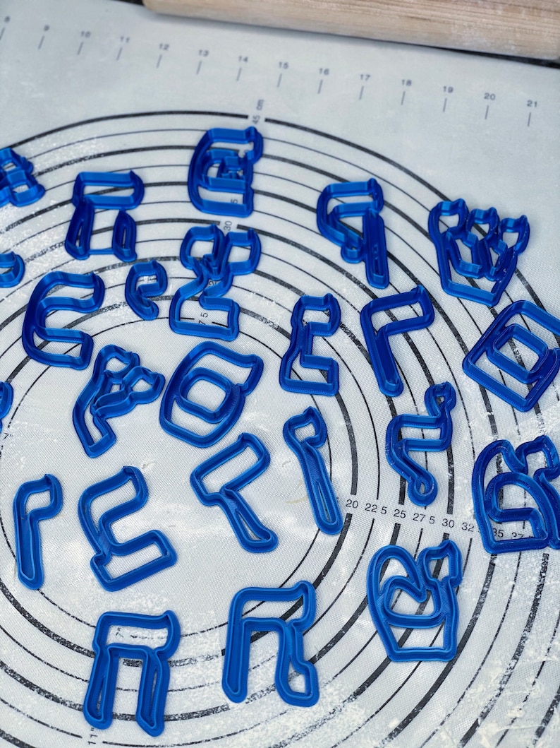 May include: A set of blue plastic cookie cutters shaped like Hebrew letters. The cutters are arranged in a circle on a white surface with a grid pattern.