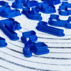 May include: A set of blue plastic cookie cutters in various shapes, including letters and swirls, on a white surface with black lines.