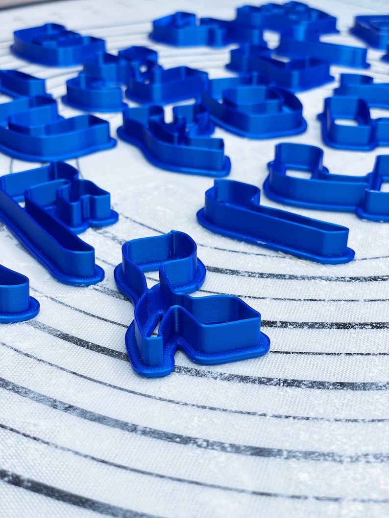 May include: A set of blue plastic cookie cutters in various shapes, including letters and swirls, on a white surface with black lines.