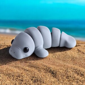 May include: A gray, rubber toy shaped like a manatee. The toy has a textured surface and black eyes. The toy is lying on a sandy beach.