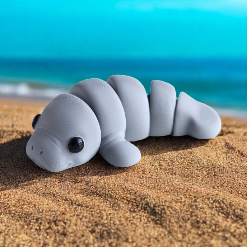 May include: A gray, rubber toy shaped like a manatee. The toy has a textured surface and black eyes. The toy is lying on a sandy beach.