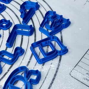 May include: A set of blue plastic cookie cutters in various shapes, including a letter, a number, and a geometric shape. The cutters are on a white, lightly floured surface.