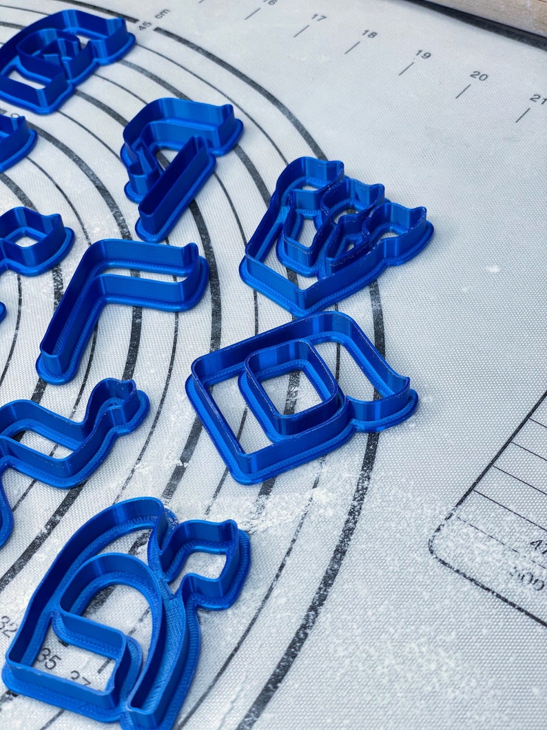 May include: A set of blue plastic cookie cutters in various shapes, including a letter, a number, and a geometric shape. The cutters are on a white, lightly floured surface.