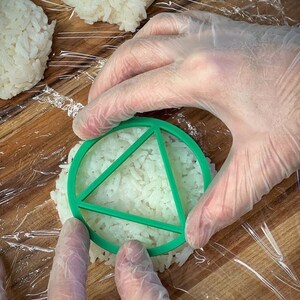 May include: A gloved hand presses a green plastic mold into a mound of white rice. The mold has a triangular shape.