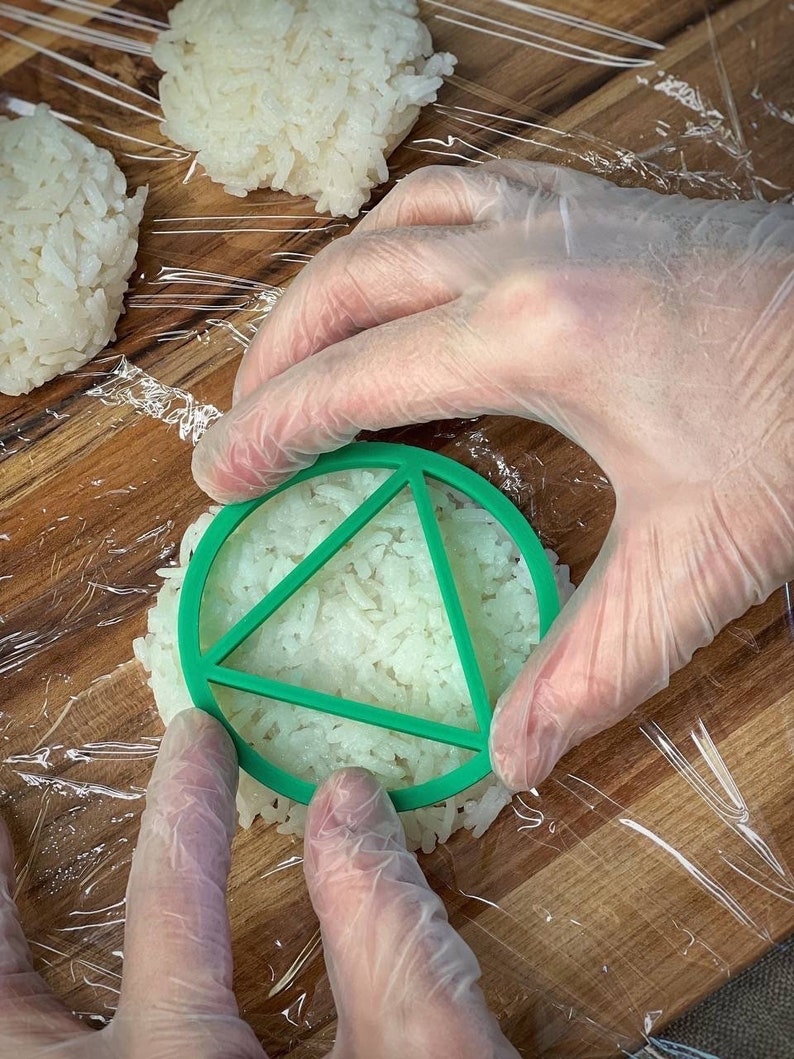 May include: A gloved hand presses a green plastic mold into a mound of white rice. The mold has a triangular shape.