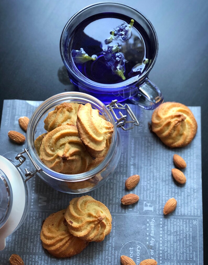 Puede incluir: Un tarro de vidrio lleno de galletas de almendras, con una taza de t&eacute; azul y almendras esparcidas sobre una superficie con estampado gris y blanco.