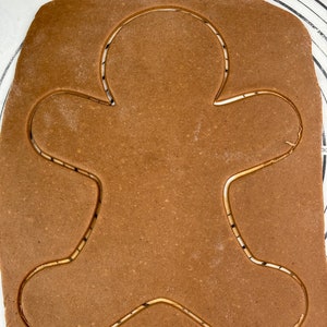 May include: Brown gingerbread cookie dough rolled out on a white surface with a gingerbread man shape cut out.