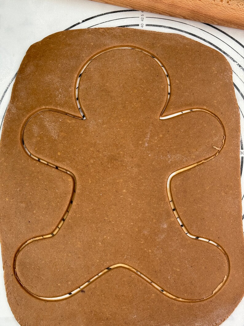 May include: Brown gingerbread cookie dough rolled out on a white surface with a gingerbread man shape cut out.