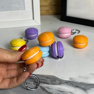 May include: A set of colorful macaron-shaped keychains. The keychains are made of plastic and feature different colors, including yellow, orange, pink, purple, and blue. Each macaron has a white filling and a silver keychain ring.