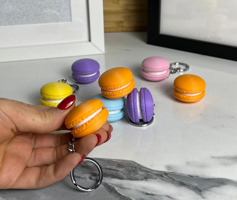 May include: A set of colorful macaron-shaped keychains. The keychains are made of plastic and feature different colors, including yellow, orange, pink, purple, and blue. Each macaron has a white filling and a silver keychain ring.