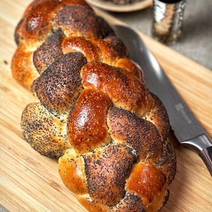 May include: A braided loaf of bread with poppy seeds on a wooden cutting board. The bread is golden brown and has a shiny glaze.