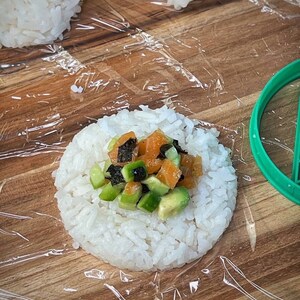 May include: A close-up of a white rice ball topped with diced avocado, salmon, and seaweed. The rice ball is on a wooden surface covered in plastic wrap.