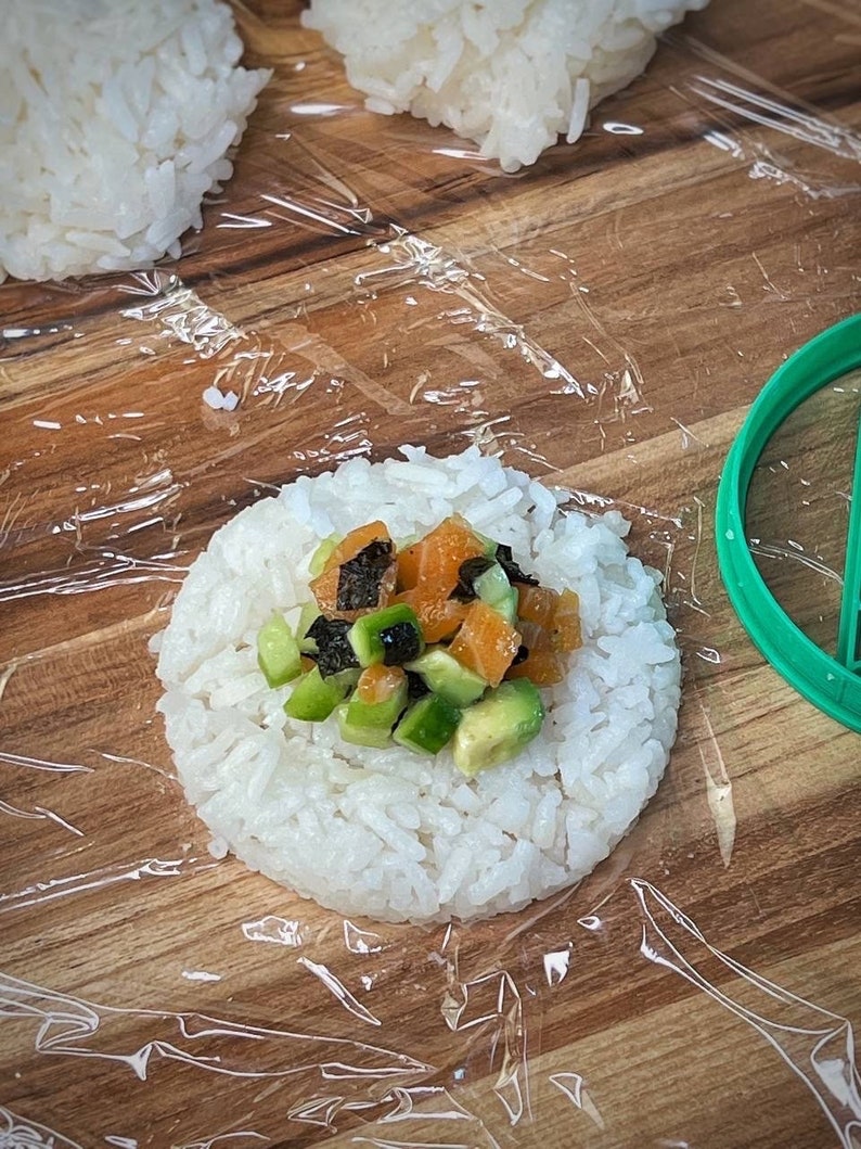 May include: A close-up of a white rice ball topped with diced avocado, salmon, and seaweed. The rice ball is on a wooden surface covered in plastic wrap.