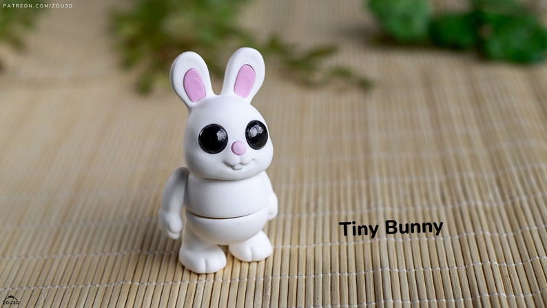 May include: A white ceramic bunny figurine with pink ears and large black eyes. The bunny is standing on a woven bamboo mat. The text "Tiny Bunny" is written below the bunny.