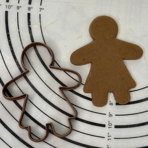 May include: A brown gingerbread person cookie cutout on a white baking mat with a brown metal gingerbread person cookie cutter. The baking mat has inch markings.