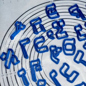 May include: A set of blue plastic cookie cutters shaped like letters of the alphabet. The cutters are arranged in a circle on a white surface with a grid pattern.