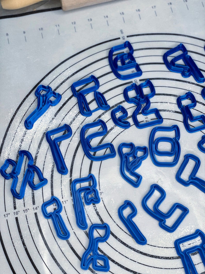 May include: A set of blue plastic cookie cutters shaped like letters of the alphabet. The cutters are arranged in a circle on a white surface with a grid pattern.