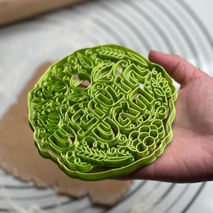 May include: Green plastic cookie cutter with a detailed design featuring the words "Be Kind" surrounded by leaves and vines.