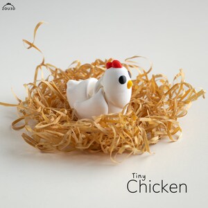 Può includere: Una piccola statuina di pollo bianca con dettagli rossi e neri si trova in un nido di carta triturata. La statuina è etichettata "Tiny Chicken".