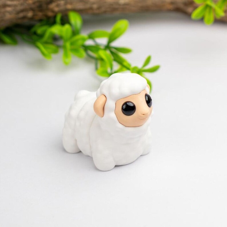 Puede incluir: Una peque&ntilde;a figura de oveja blanca con un cuerpo texturizado y esponjoso y una cara color canela. La oveja tiene ojos grandes y negros y orejas peque&ntilde;as y color canela. La figura est&aacute; sobre una superficie blanca, con follaje verde en el fondo.
