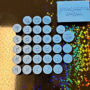 May include: A selection of light blue 3D-printed Hanukkah-themed items. The image shows small, circular tokens with diverse symbols and Hebrew letters, plus a rectangular piece with Hebrew text. The tokens feature a menorah, Star of David, and other designs.