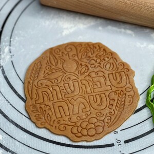 May include: A brown cookie dough circle with a decorative design and the Hebrew words "Shanah Tovah" in the centre. The dough is on a white silicone baking mat with a circular pattern.