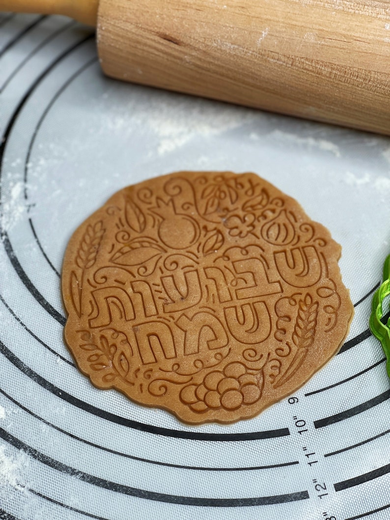 May include: A brown cookie dough circle with a decorative design and the Hebrew words "Shanah Tovah" in the centre. The dough is on a white silicone baking mat with a circular pattern.