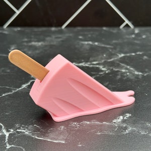 Puede incluir: Un helado de paleta rosa con un palo de madera, derriti&eacute;ndose sobre una superficie de m&aacute;rmol negro.