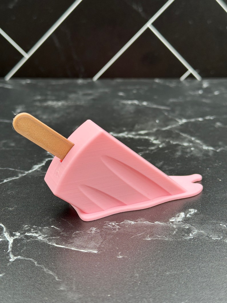 Puede incluir: Un helado de paleta rosa con un palo de madera, derriti&eacute;ndose sobre una superficie de m&aacute;rmol negro.