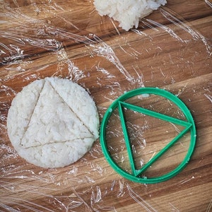 May include: A green plastic mold shaped like a circle with a triangle inside. The mold is on a wooden surface with a white rice patty inside.