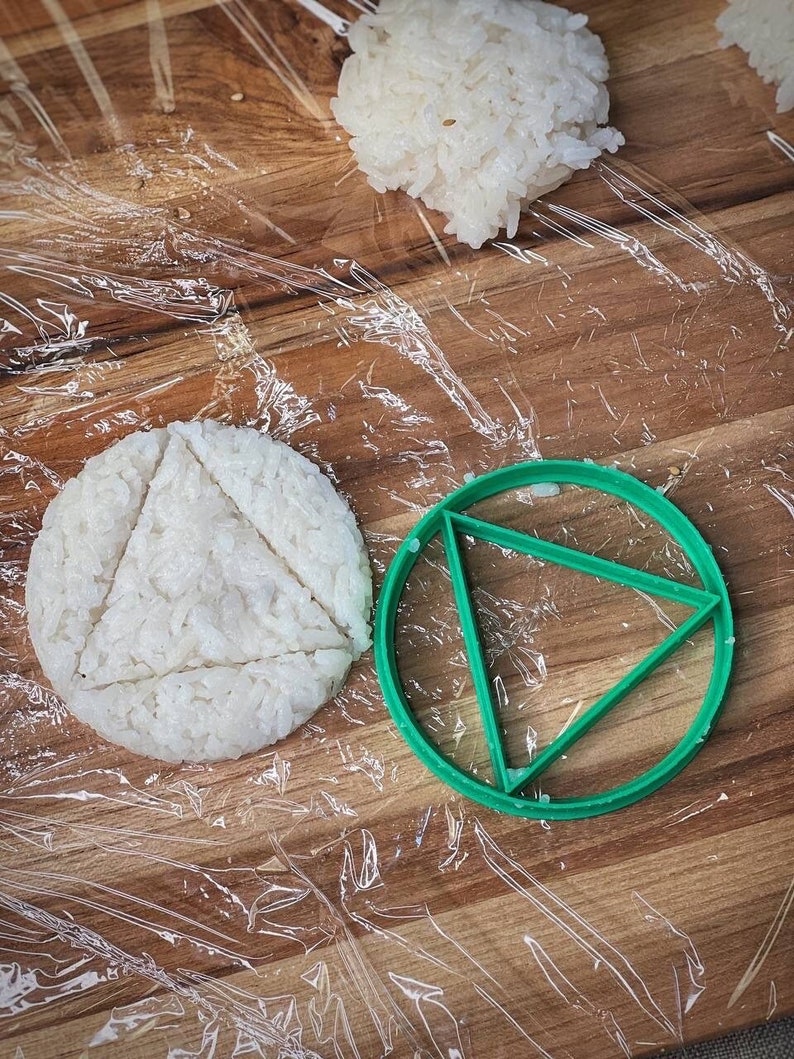 May include: A green plastic mold shaped like a circle with a triangle inside. The mold is on a wooden surface with a white rice patty inside.