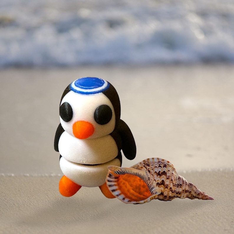 May include: A white penguin figurine with a blue and white hat, orange beak, and black wings, standing on a sandy beach next to a brown seashell.