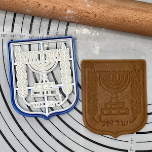 Puede incluir: Un cortador de galletas de plástico blanco con forma del escudo de armas de Israel. El cortador está sobre una estera de hornear de silicona blanca y azul. Una galleta marrón con el mismo diseño está al lado del cortador. La galleta tiene la palabra "ישראל" escrita en ella.