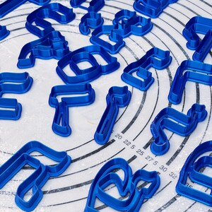 May include: A set of blue plastic cookie cutters in various shapes, including letters and abstract designs. The cutters are arranged on a white surface with a circular pattern.