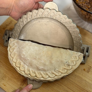 May include: A silver-coloured empanada press with a decorative edge, partially open, revealing a filled pastry. The pastry is light brown and has a braided edge. The press is on a wooden chopping board.