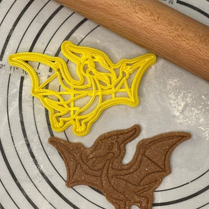 Peut inclure: Deux emportes-pièces en forme de dinosaure, l'un jaune et l'autre brun, sur une surface à motifs blancs et jaunes. L'emporte-pièce jaune est un ptérodactyle et l'emporte-pièce brun est un dinosaure.