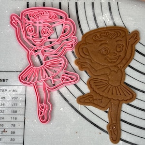 Könnte beinhalten: Ein rosa Kunststoff-Ausstecher in Form einer Ballerina, neben einem gebackenen Lebkuchen-Keks in der gleichen Form. Der Ausstecher befindet sich auf einer weiß-grauen Oberfläche mit Zollmaßen.