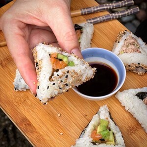 May include: A close-up of a hand holding a triangular sushi roll with salmon, avocado, and sesame seeds. The roll is on a wooden cutting board with other sushi rolls and a small bowl of soy sauce.