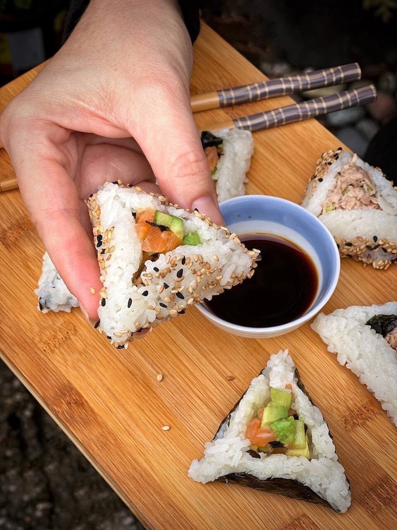May include: A close-up of a hand holding a triangular sushi roll with salmon, avocado, and sesame seeds. The roll is on a wooden cutting board with other sushi rolls and a small bowl of soy sauce.