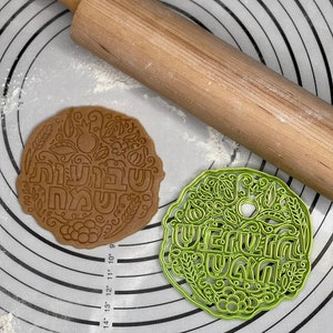 May include: Two cookie cutters, one brown and one green, with Hebrew lettering and floral designs. The cutters are on a white silicone baking mat with concentric circles.