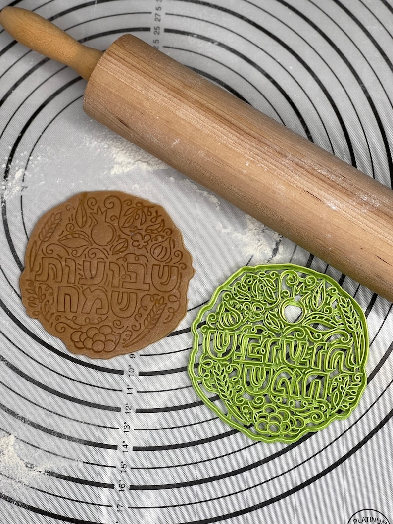 May include: Two cookie cutters, one brown and one green, with Hebrew lettering and floral designs. The cutters are on a white silicone baking mat with concentric circles.